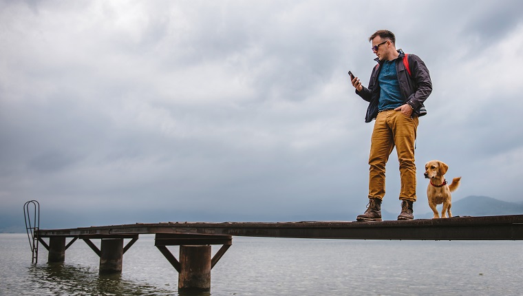 Man standing on wooden dock on stormy weather with his dog and using smart phone. He is wearing red backpack, blue jacket and leather boots